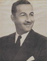A black-and-white portrait of a smiling person wearing a suit, tie, and shirt, looking slightly toward the right.