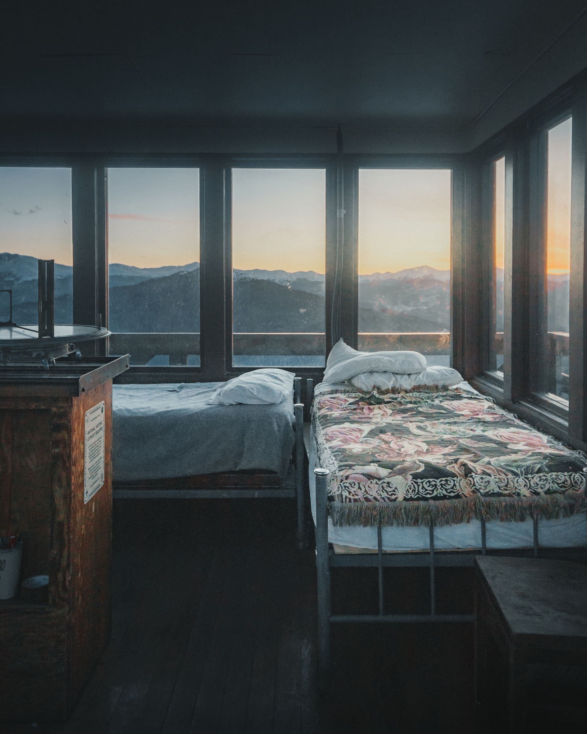 Two beds by tall windows with a mountain sunset view, warm light filling a cozy room.