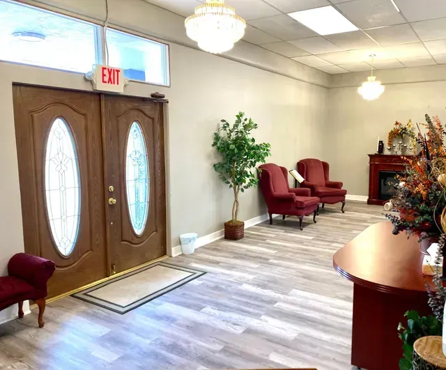 Bright lobby with double wooden doors, red chairs, plants, chandelier, and reception desk.