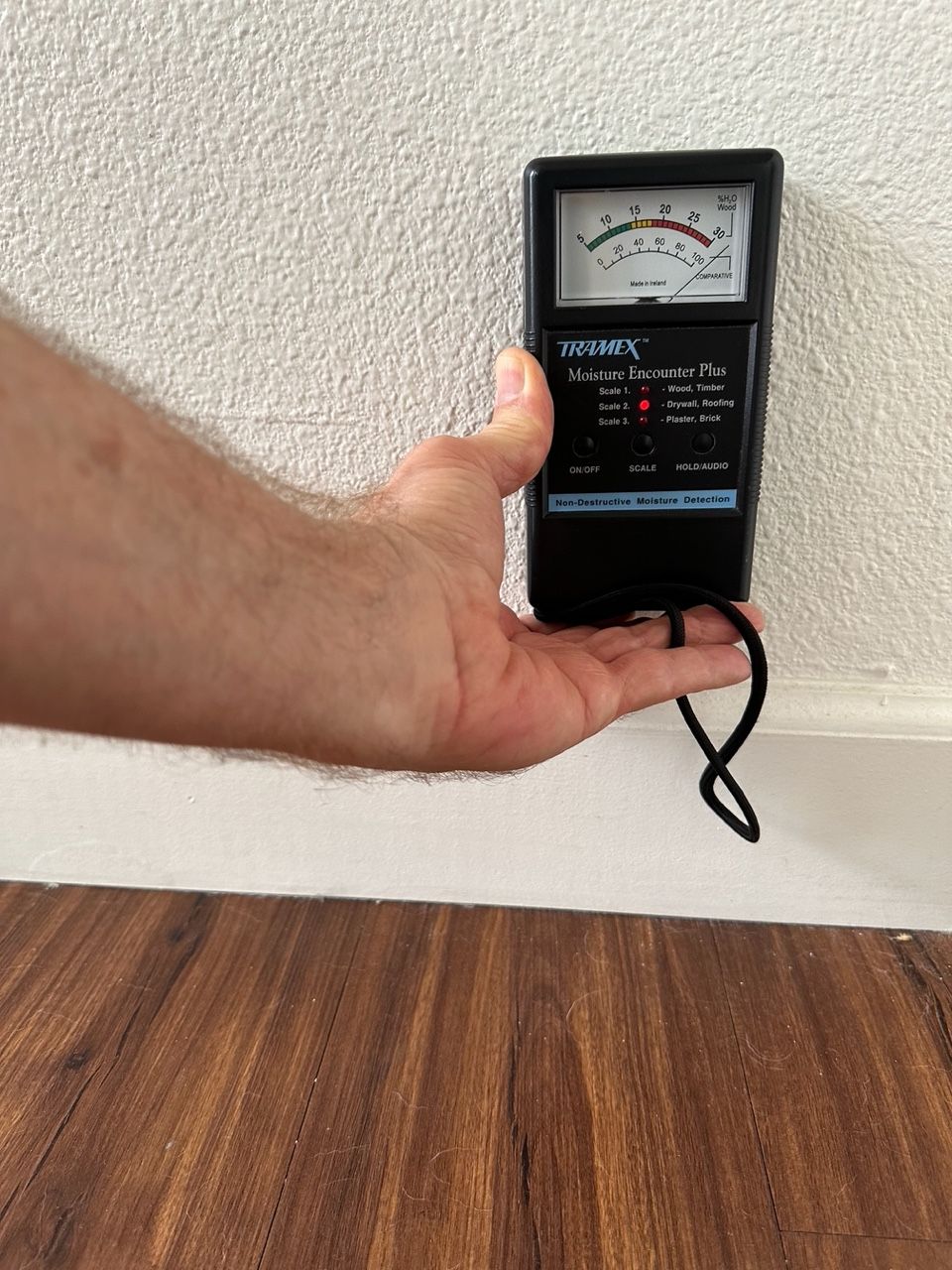 Hand holding a black moisture meter against a white wall and a brown floor. Meter reads near zero.