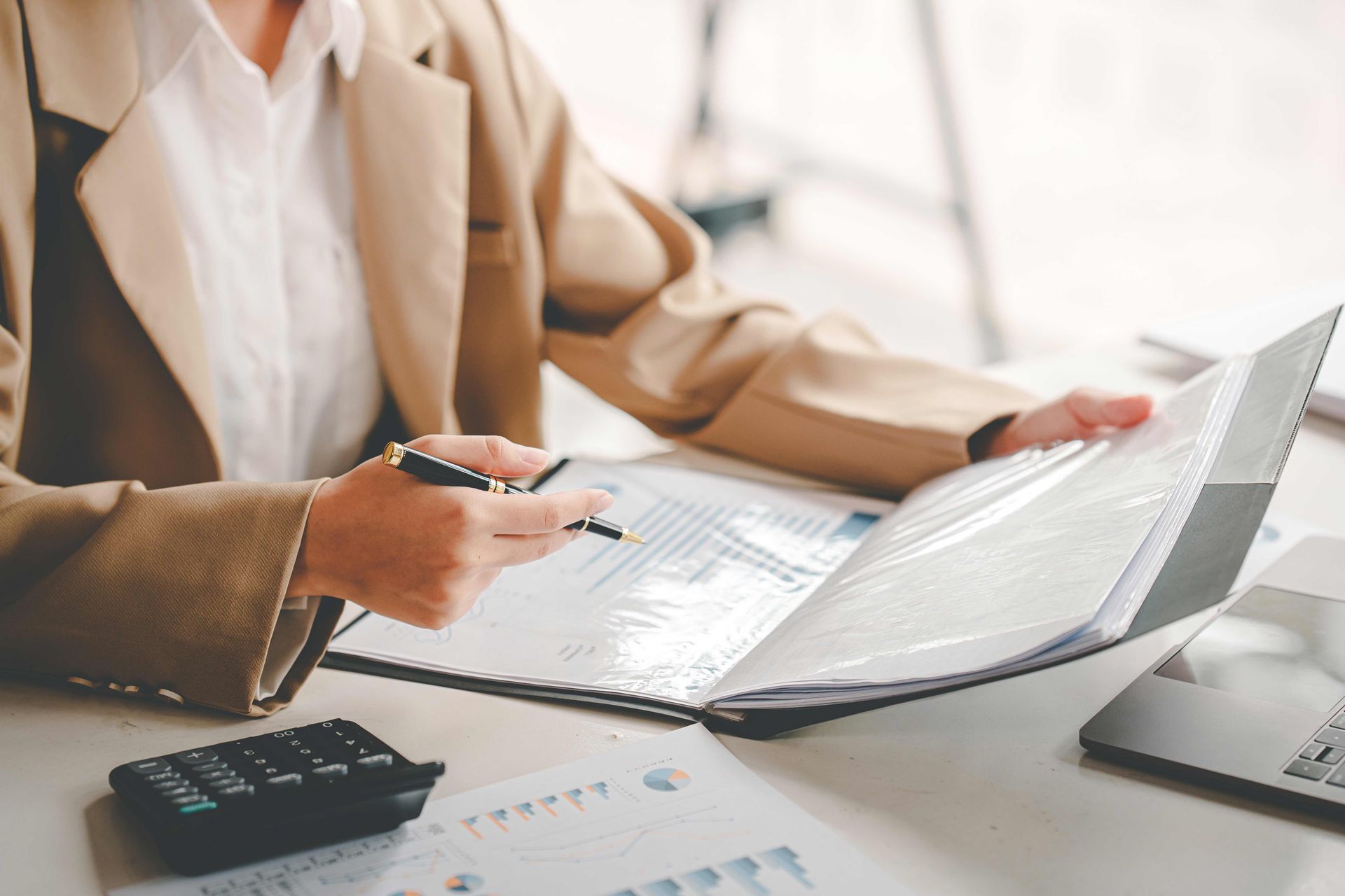 Businesswoman reviewing financial charts and reports in binder with calculator and laptop on desk.