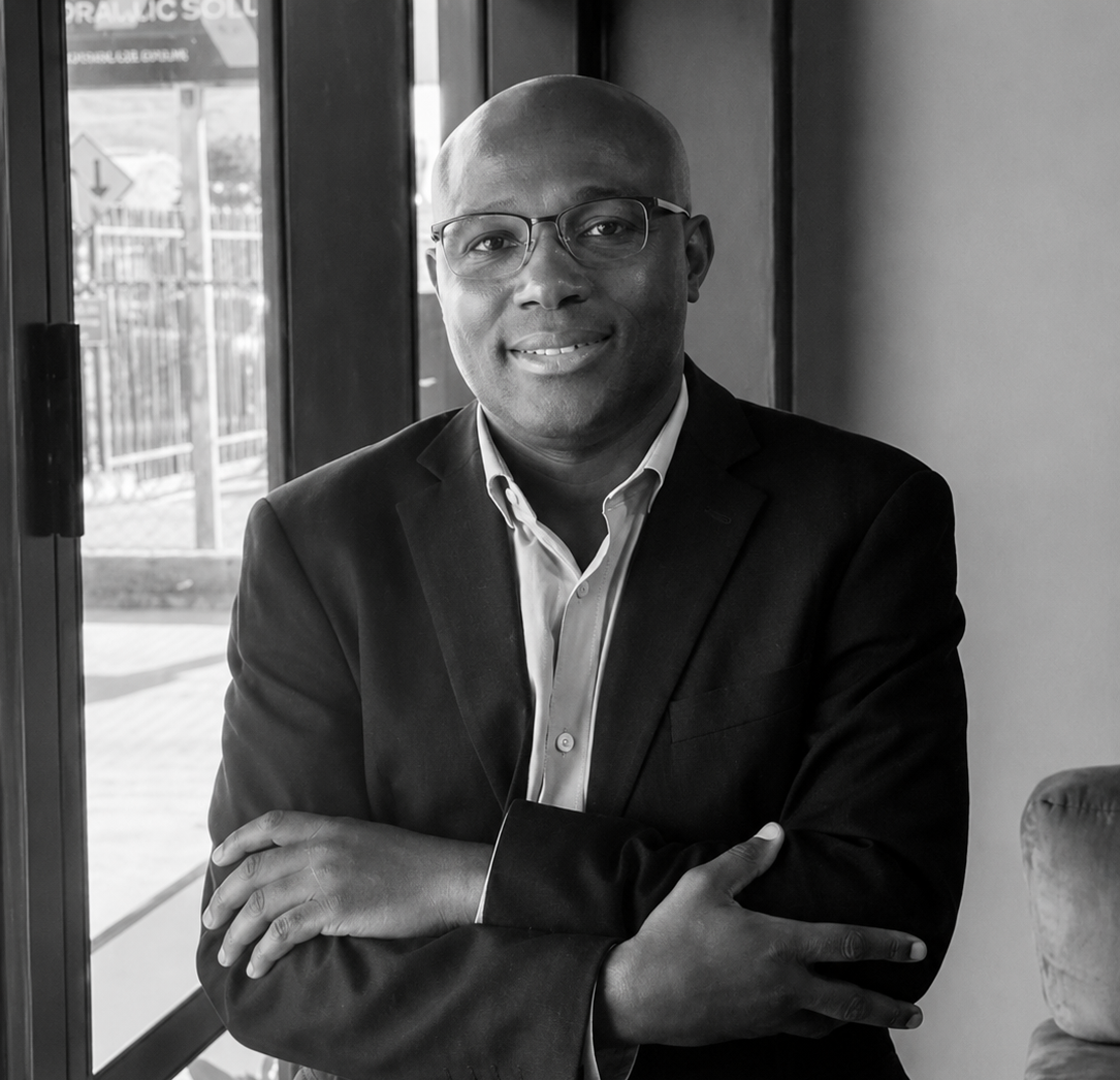 Titus (man) smiling at camera in glasses and blazer standing with arms crossed by a window.