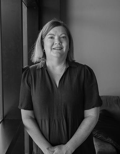 Carolyn (woman) standing indoors beside a window, smiling gently in a dark dress.