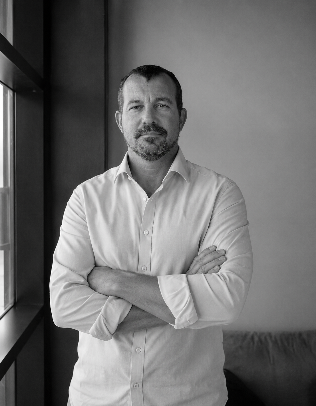Stewart (man) standing with arms crossed in a white shirt beside a window in a simple room.