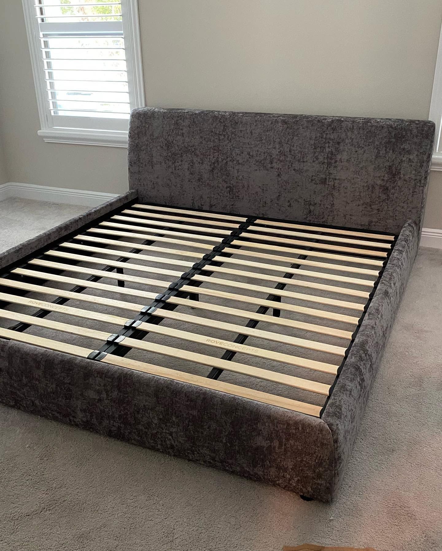 Gray upholstered bed frame with slatted base in a carpeted room near a window.