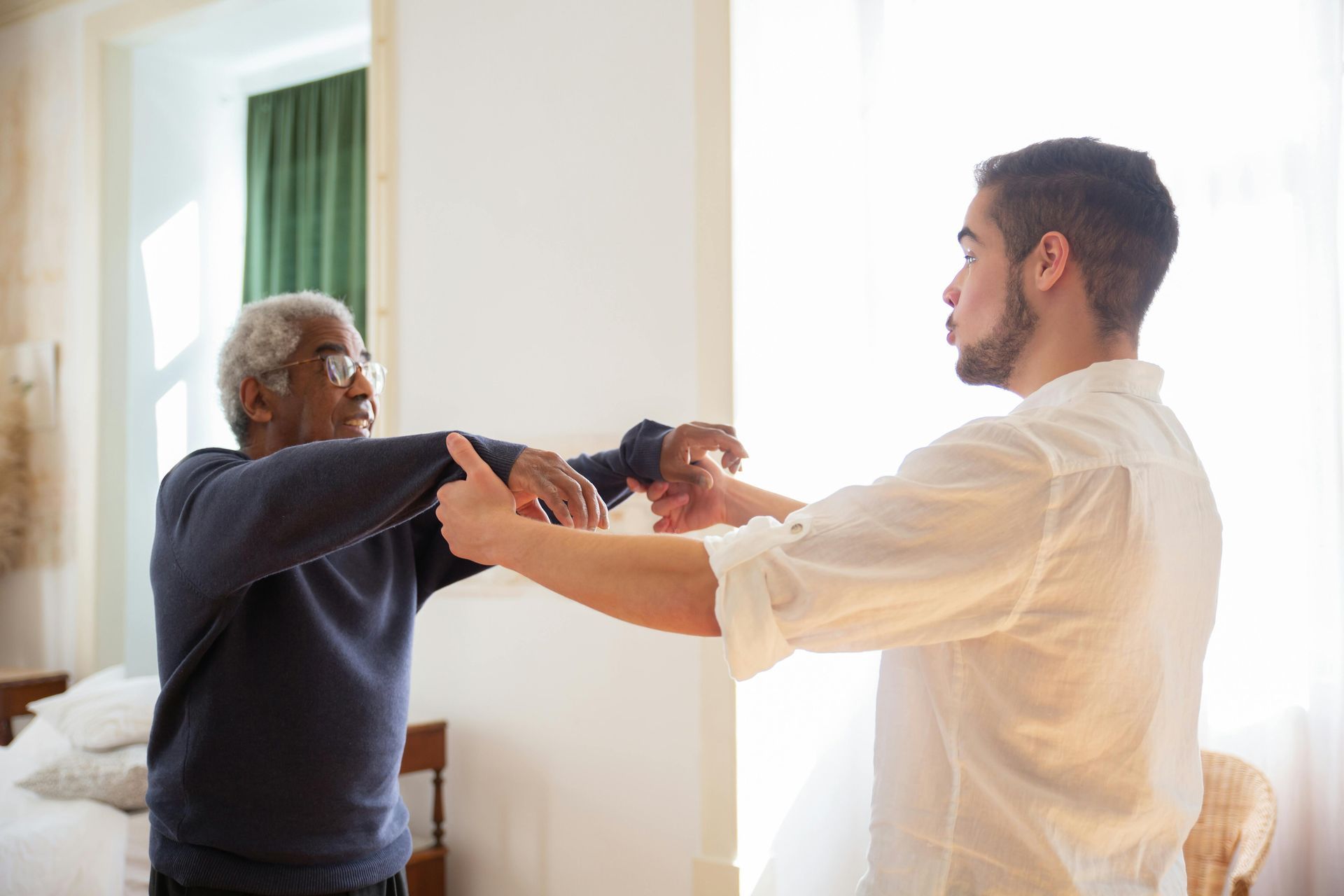 Man assisting another with arm exercises indoors, near a bed and window.