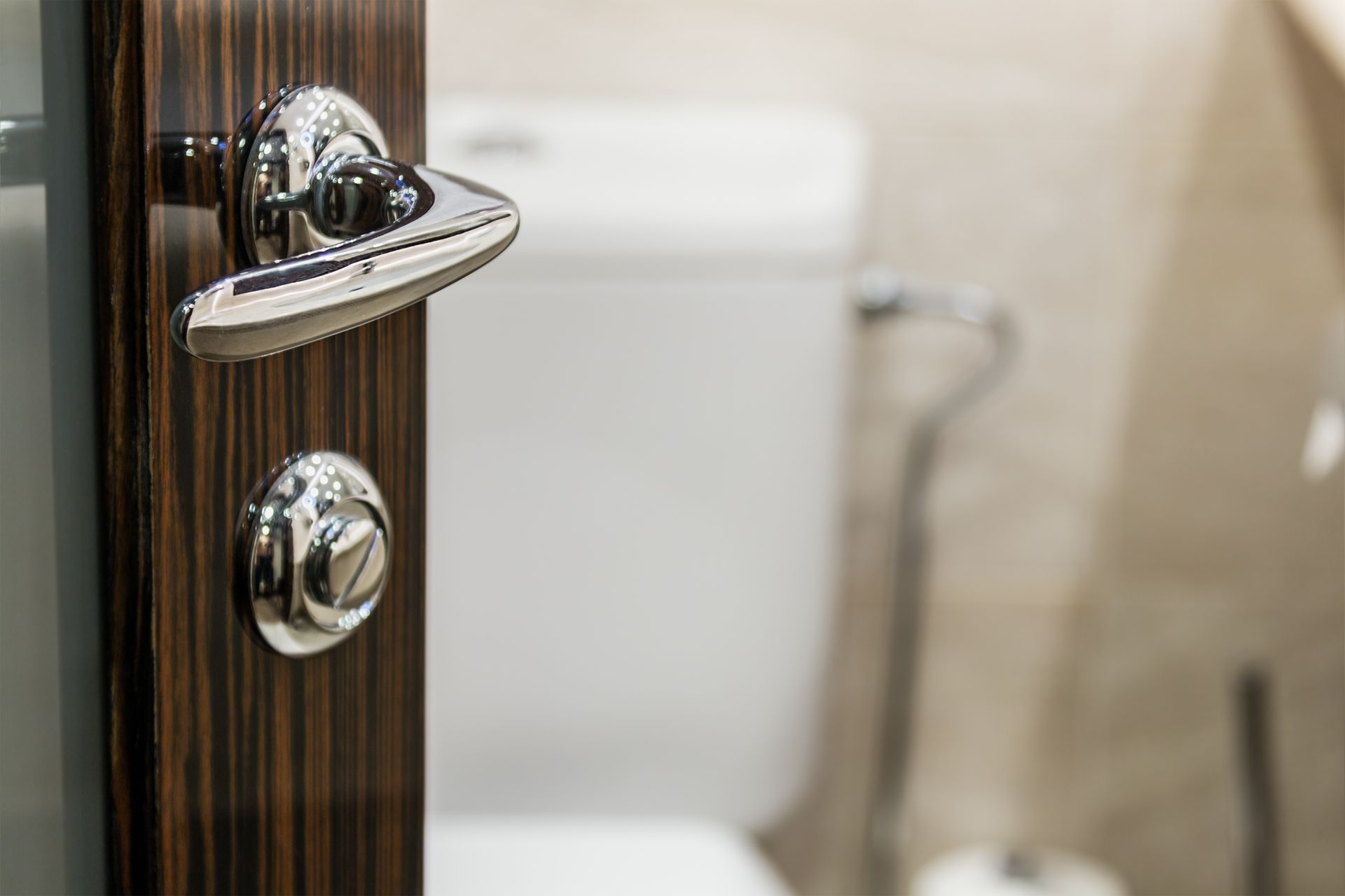 Chrome door handle and lock on a dark wood bathroom door, with a toilet visible in the background