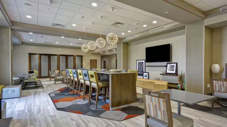 Hotel lobby with long bar, seating area, and large TV on the wall. Modern decor with colorful accents.