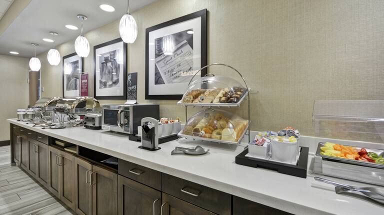 Hotel breakfast buffet with food and appliances on a counter with cabinets below, art on the wall.