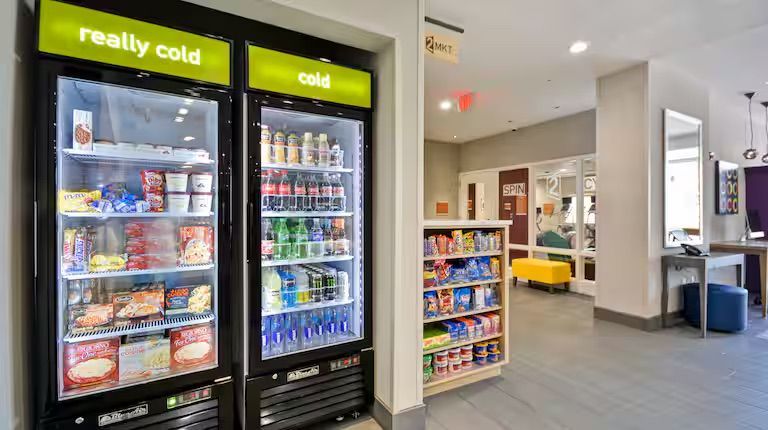 Two cold food/drink refrigerators and a snack shelf in a hotel lobby with seating.