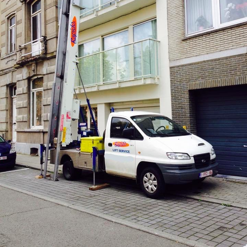 Camion blanc avec élévateur atteignant un bâtiment, stationné sur un trottoir devant un bâtiment.