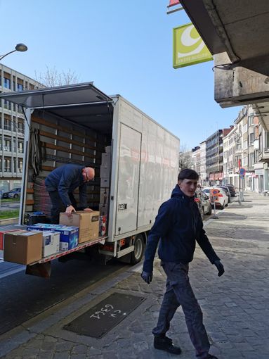 Deux hommes déchargent des cartons d'un camion garé dans une rue. Une personne passe.