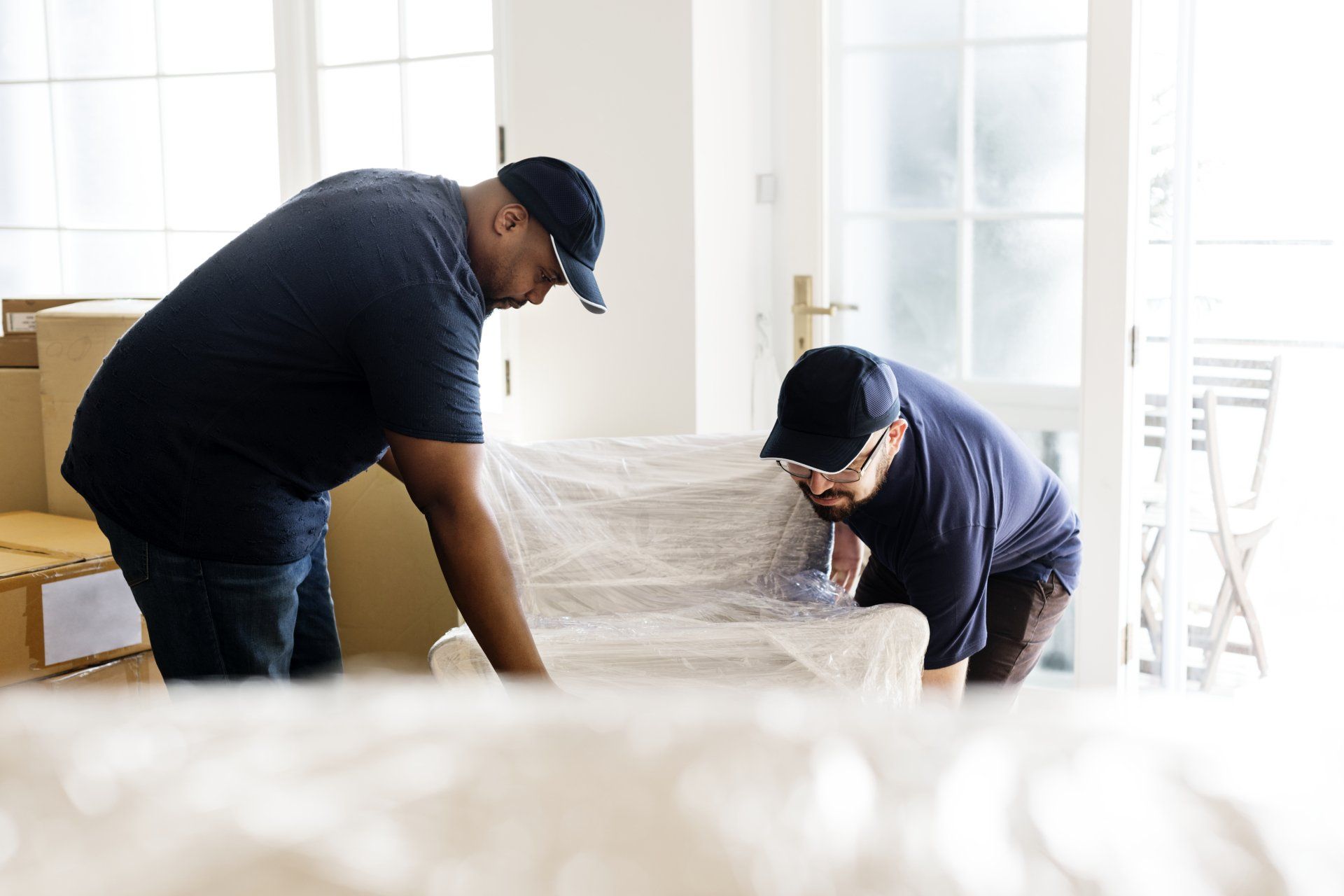 Deux déménageurs emballent une chaise dans du plastique à l'intérieur d'une maison avec des cartons et des fenêtres.