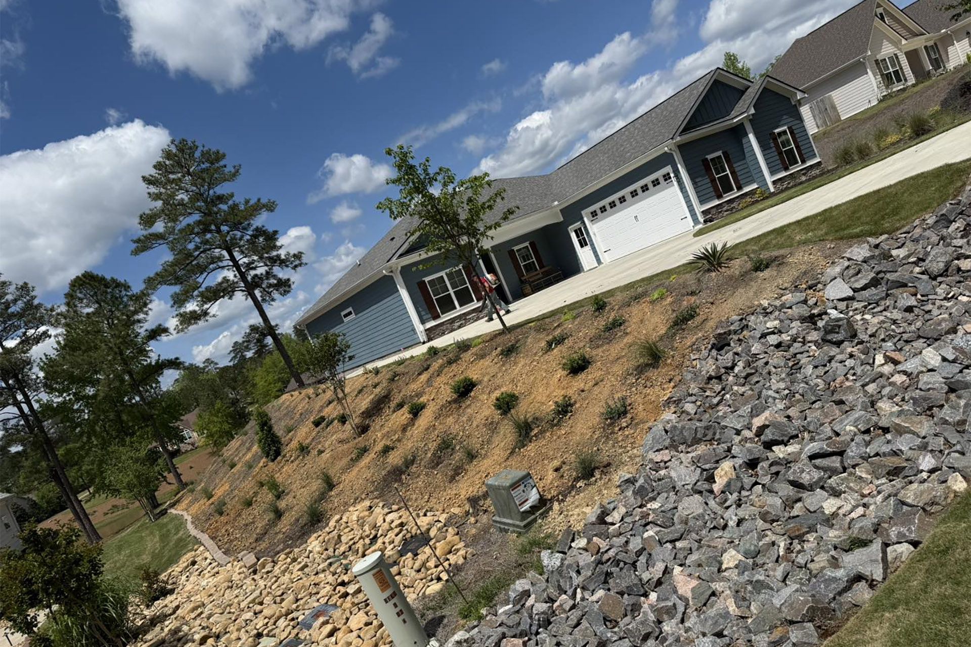 A blue suburban house sits atop a sloped, rocky landscape under a partly cloudy blue sky.