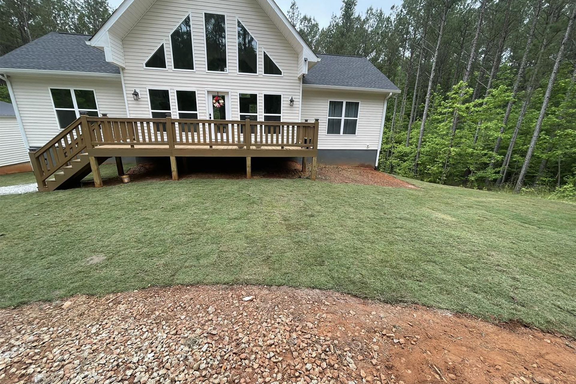 A white, A-frame house with a wooden deck and staircase sits on a grassy lawn bordered by trees and gravel.