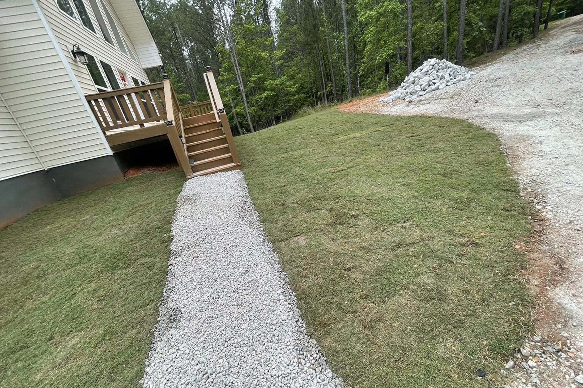 A gravel walkway leads from a gravel driveway to the wooden steps of a house deck on a grassy, sloped residential lot.