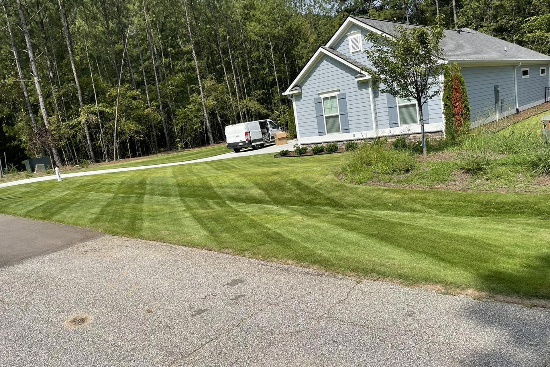 A light blue house sits on a green, freshly mowed lawn with stripe patterns, near a driveway and forest trees.