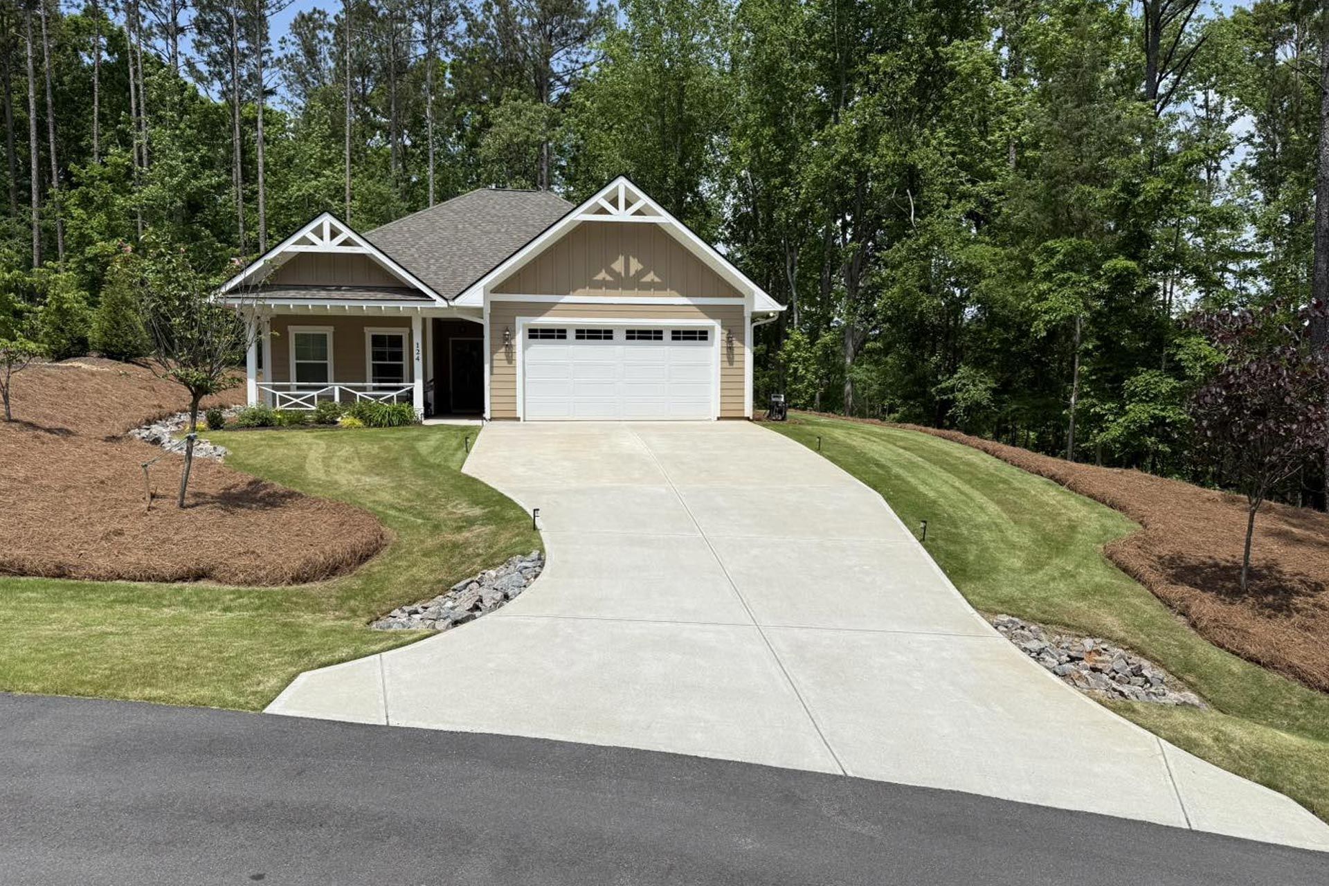 A tan, single-story house with a white garage door, a front porch, and a curved concrete driveway, set against trees.
