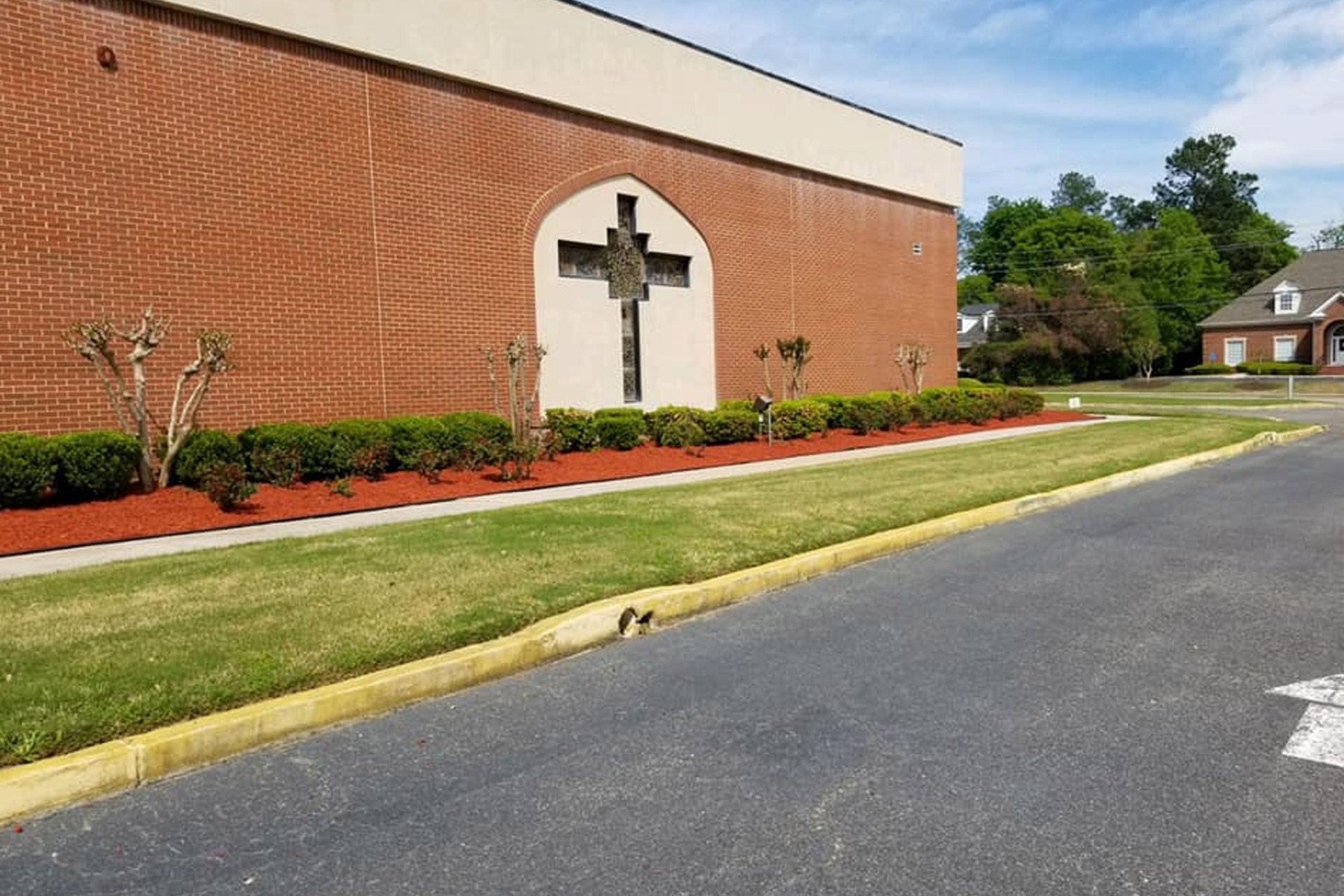 A brick church building with a large cross set in an arched inset, surrounded by landscaped bushes and a grassy lawn.