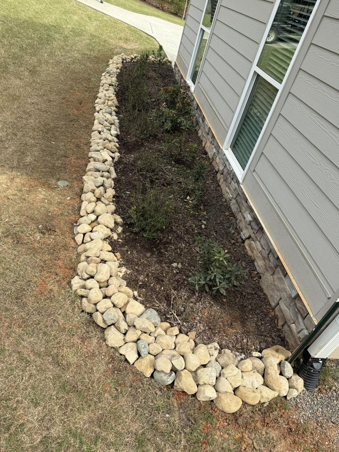 A landscaped garden bed next to a house exterior, bordered by a curved wall of rounded river stones on grass.