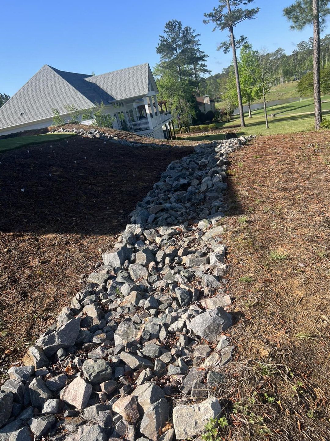 A stone-lined drainage swale runs downhill between landscaped slopes with a house in the background.