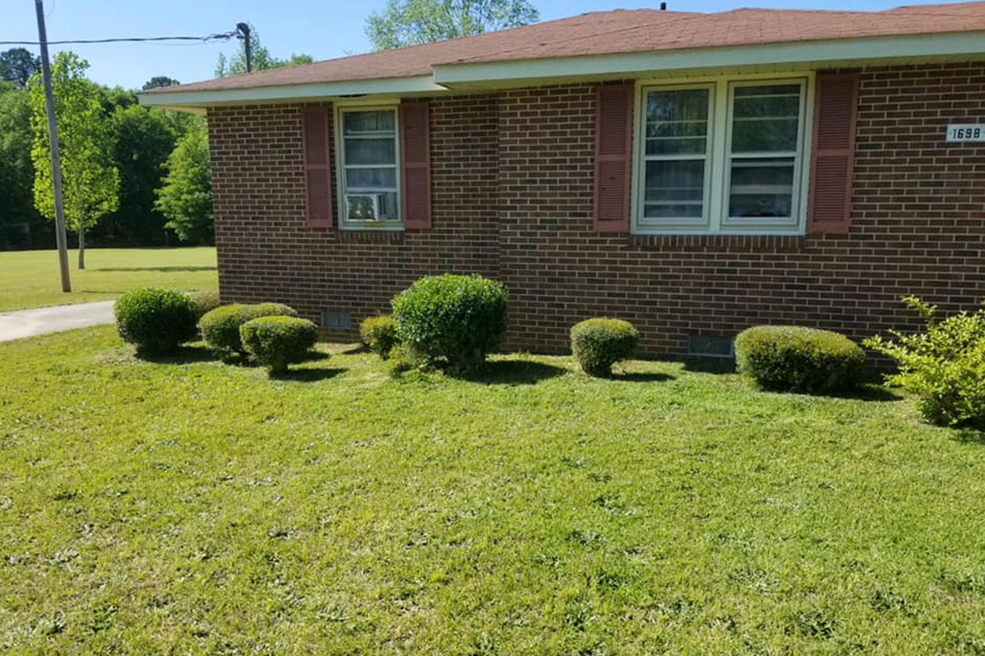 A single-story red brick house with small round shrubs in the front yard under a clear blue sky.