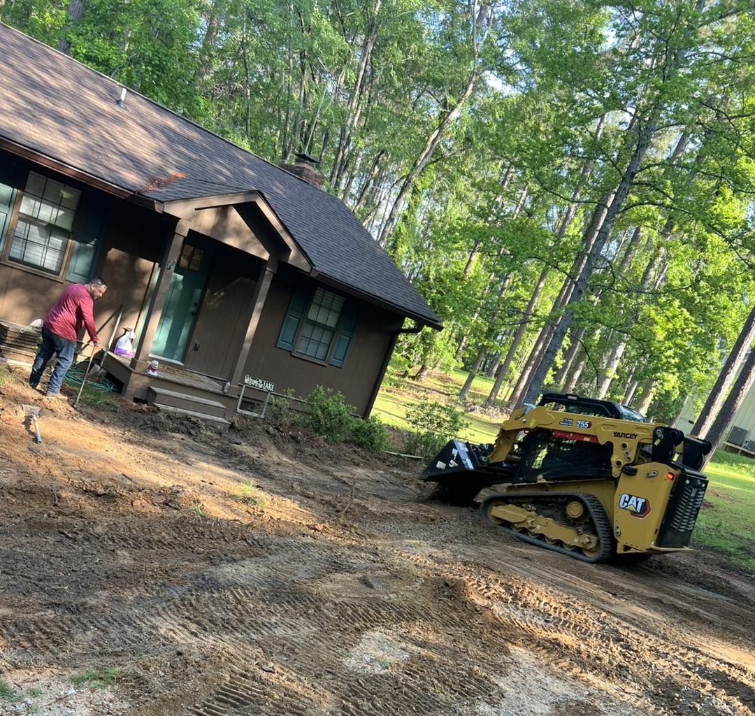 A person in a red shirt works in a dirt yard next to a brown house while a yellow Caterpillar skid steer loader operates.
