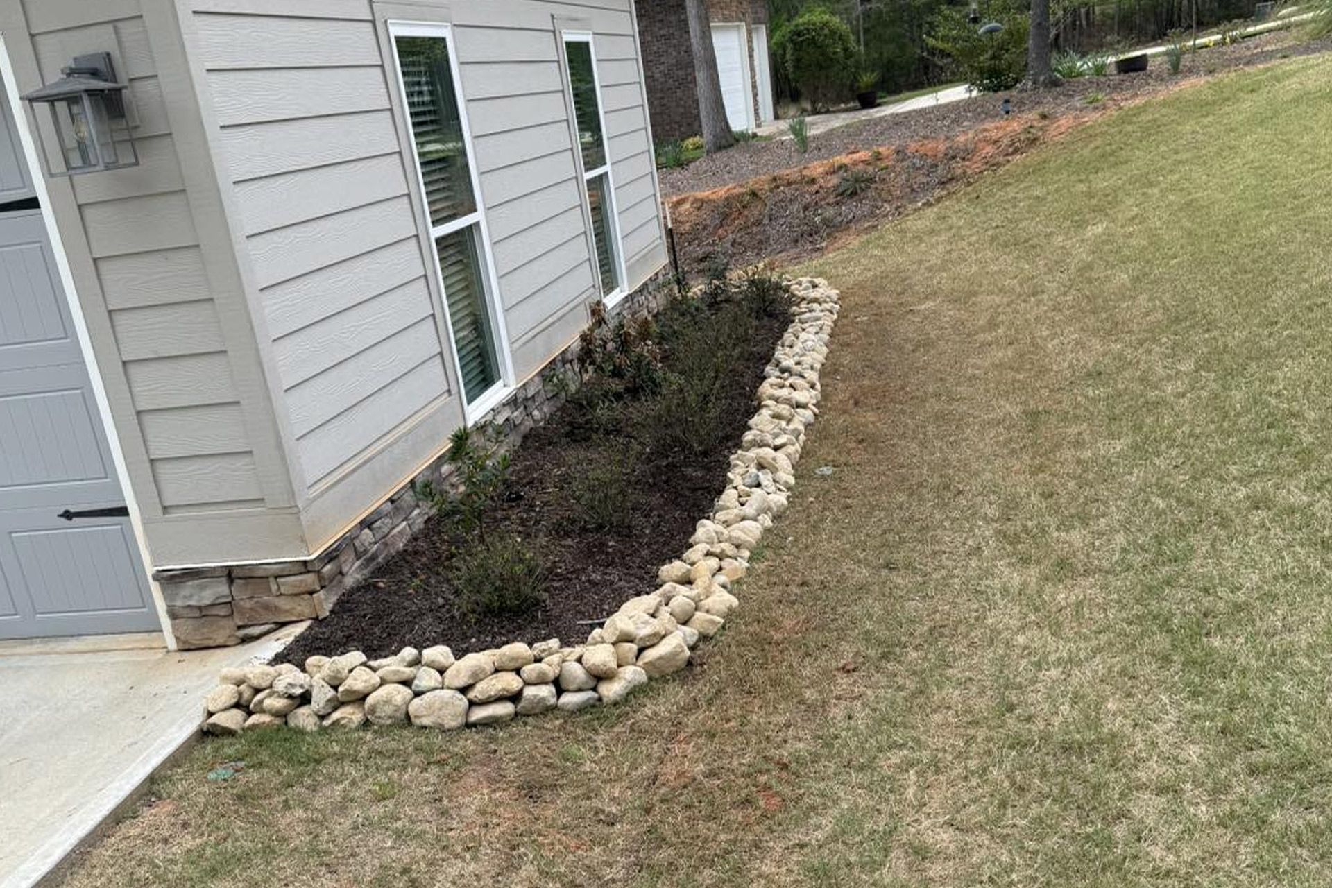 A small landscaped flower bed along the side of a house, edged with light-colored stones and filled with dark mulch.