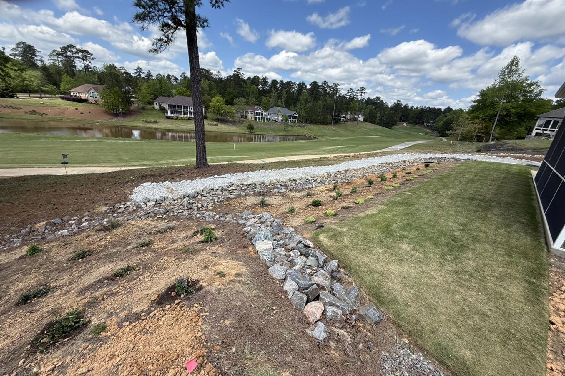 A rocky dry creek bed leads through a landscaped yard toward a pond and distant houses under a cloudy blue sky.