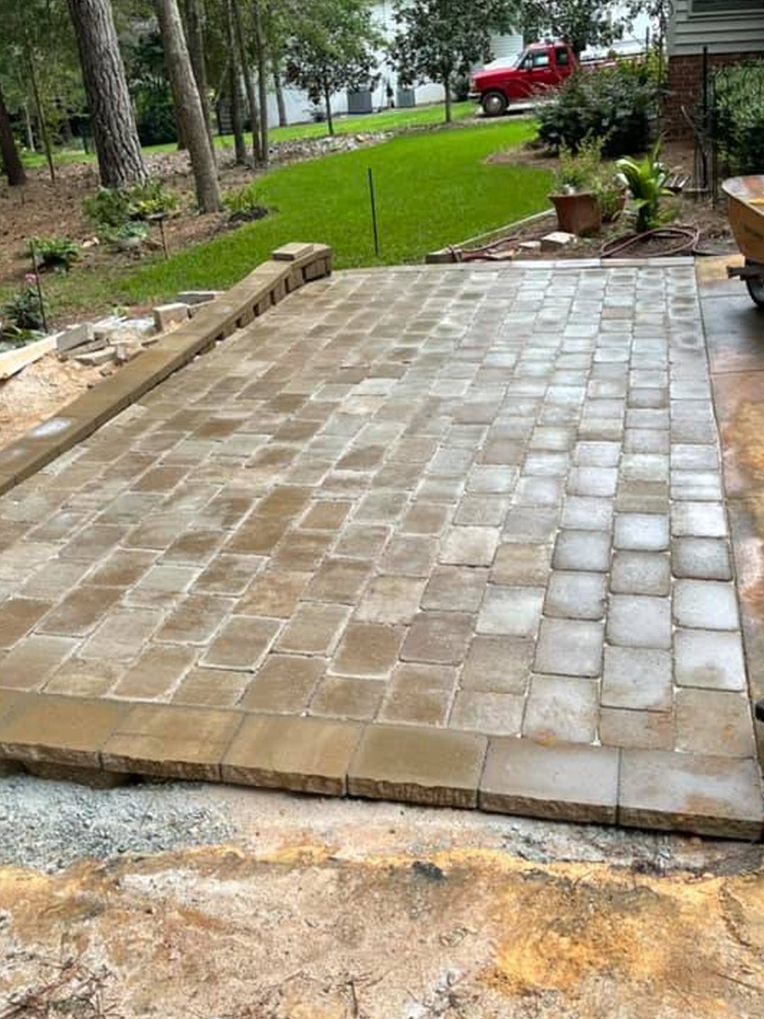 A newly installed rectangular paver patio with a raised stone border edge, set in a backyard near trees and a green lawn.
