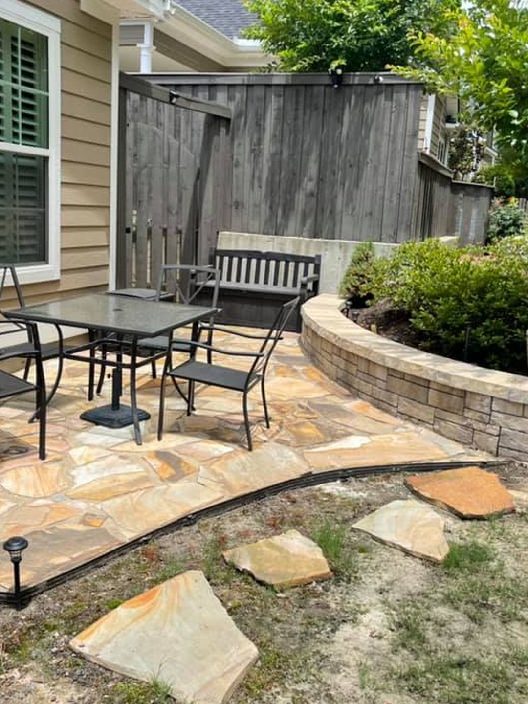 A backyard stone patio featuring a dining set with four chairs, a wooden bench, and a curved retaining wall.