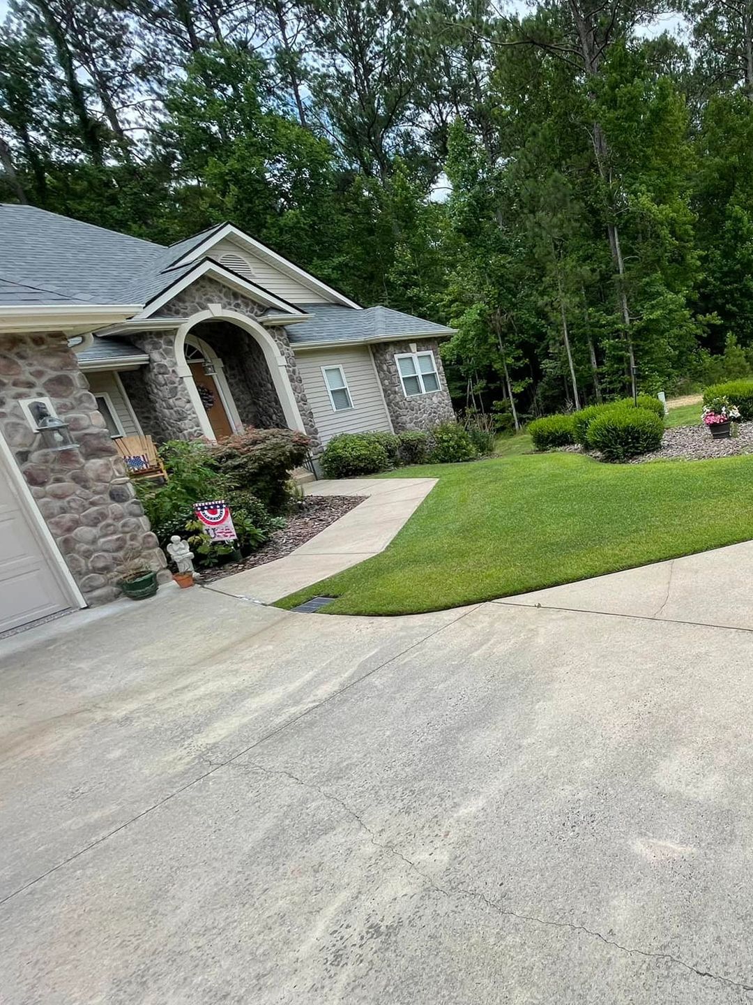 A stone-front house with a grey roof and a concrete driveway, set against a backdrop of trees and a green lawn.