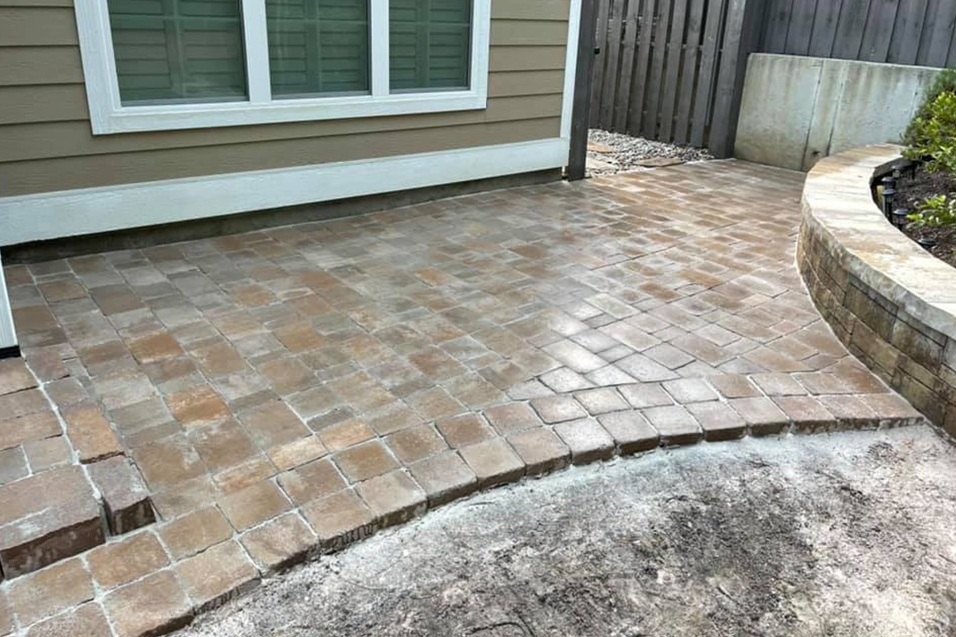 A newly installed brown paver patio sits against the side of a house next to a curved stone retaining wall.