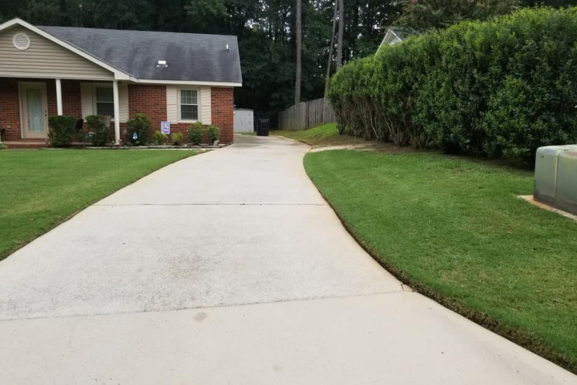 A concrete driveway leads to a brick house with a front porch, flanked by a green lawn and a tall evergreen hedge.