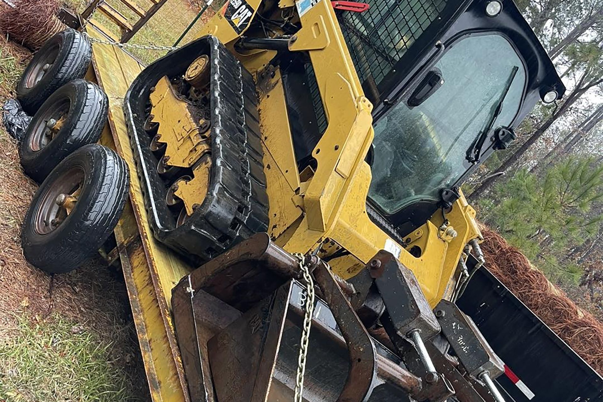 A yellow Caterpillar skid steer loader with tracks parked on a trailer in a wooded area.