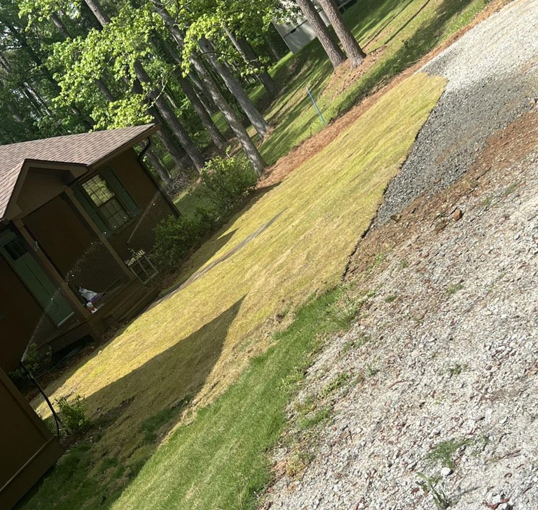 A patch of newly laid sod with exposed soil, situated next to a brown cabin and a gravel driveway in a wooded area.