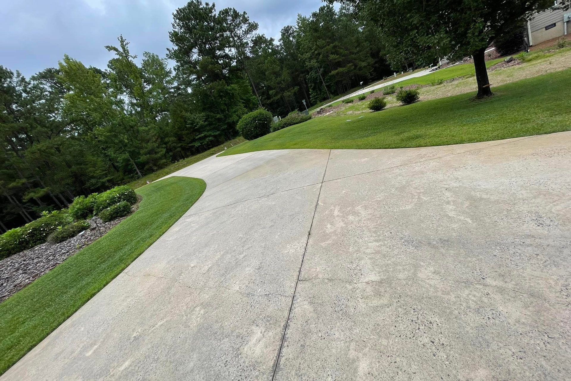 A paved driveway curves past a green lawn and landscaping, leading toward a background of trees under a cloudy sky.