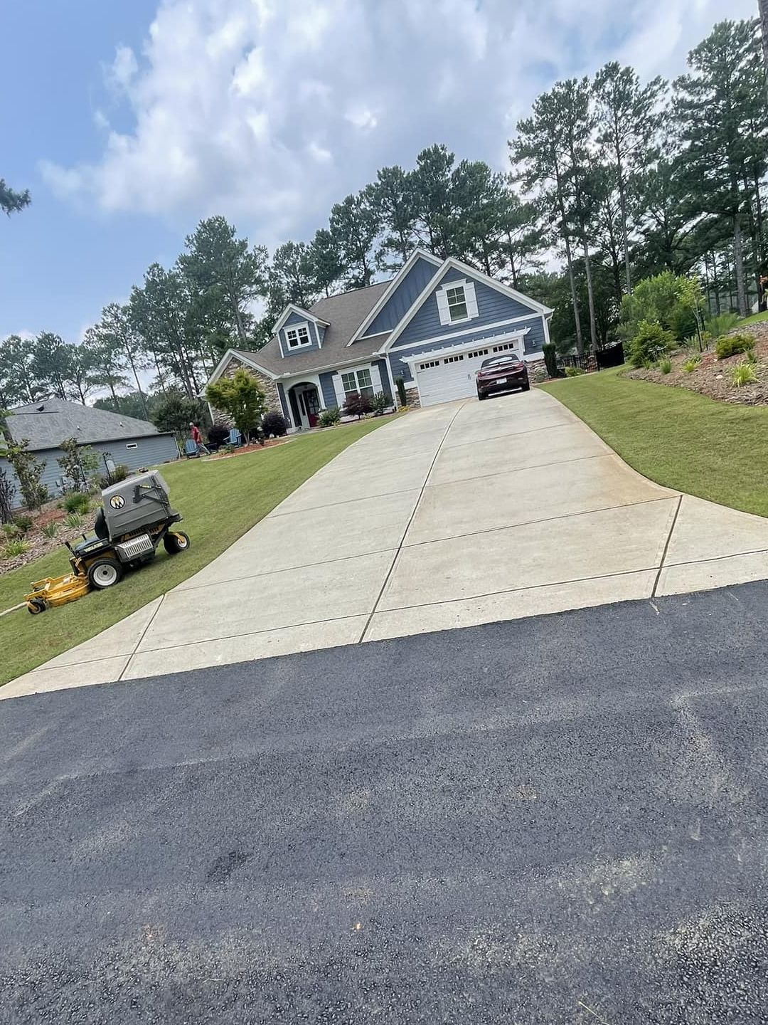 A blue suburban house with a long concrete driveway, parked car, and a riding lawnmower on the adjacent lawn.