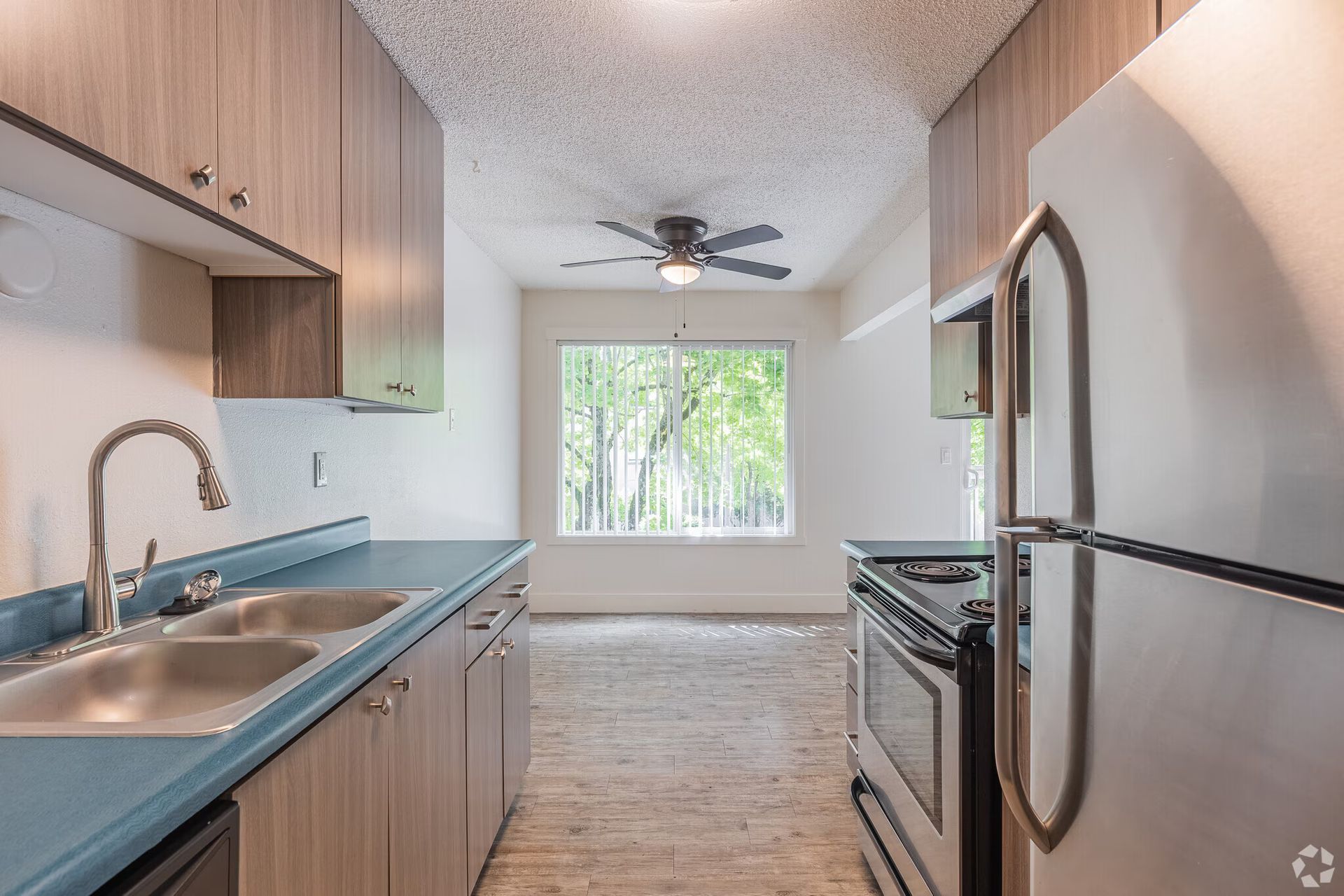 Narrow kitchen with wood cabinets, teal countertops, stainless appliances, and a window view into a living area