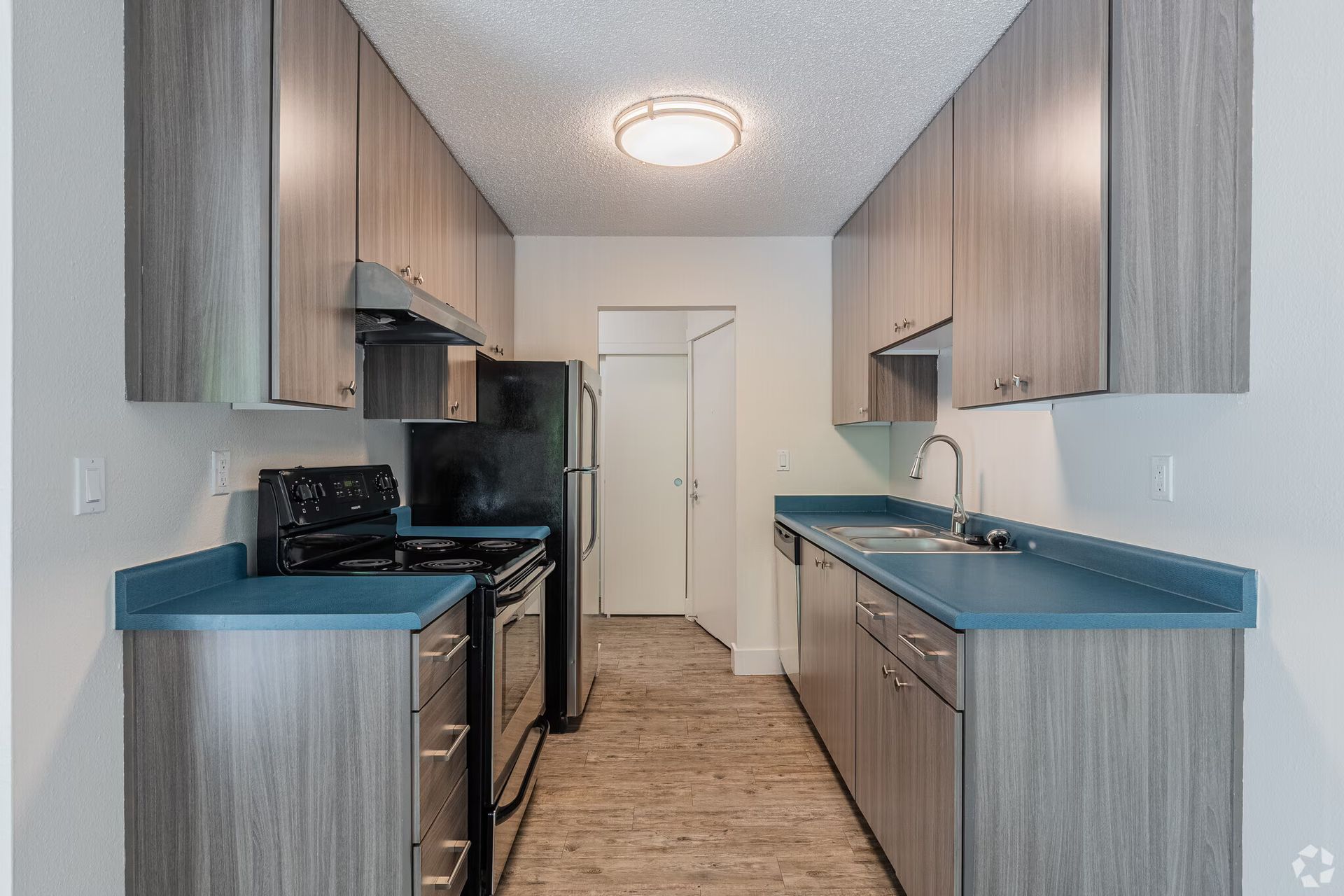 Narrow galley kitchen with wood cabinets, teal countertops, black appliances, and a white door at the end.
