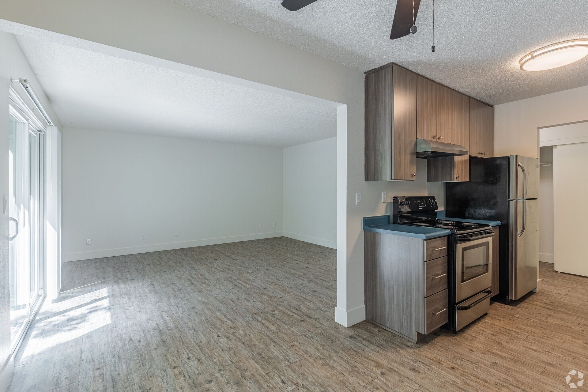 Empty living room beside a small kitchen with wood cabinets, stainless stove, and gray carpeted floors