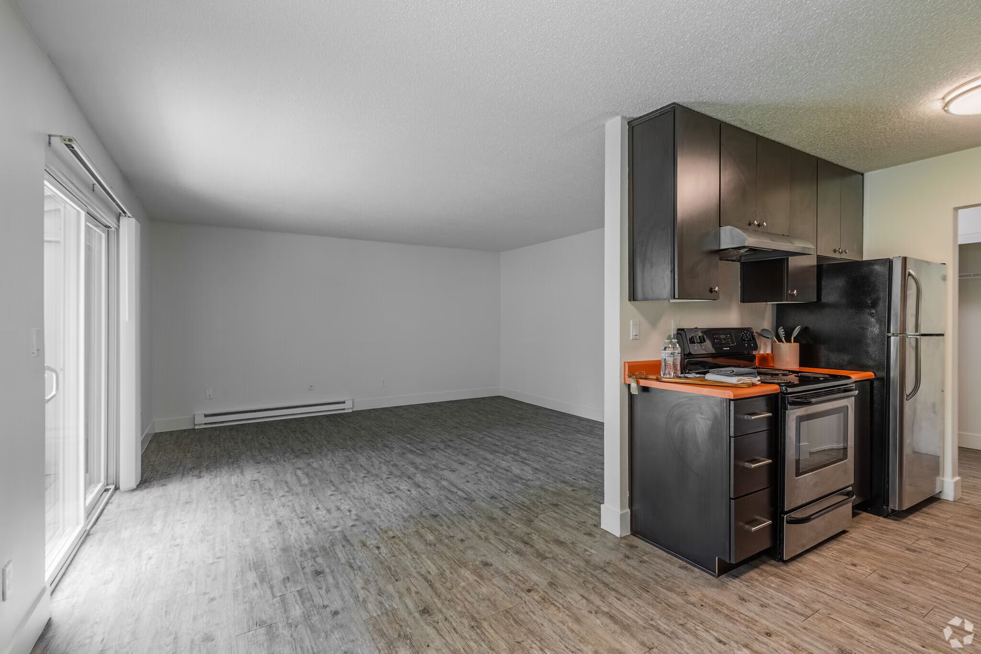 Empty living room beside a compact kitchen with gray floors, white walls, and stainless appliances