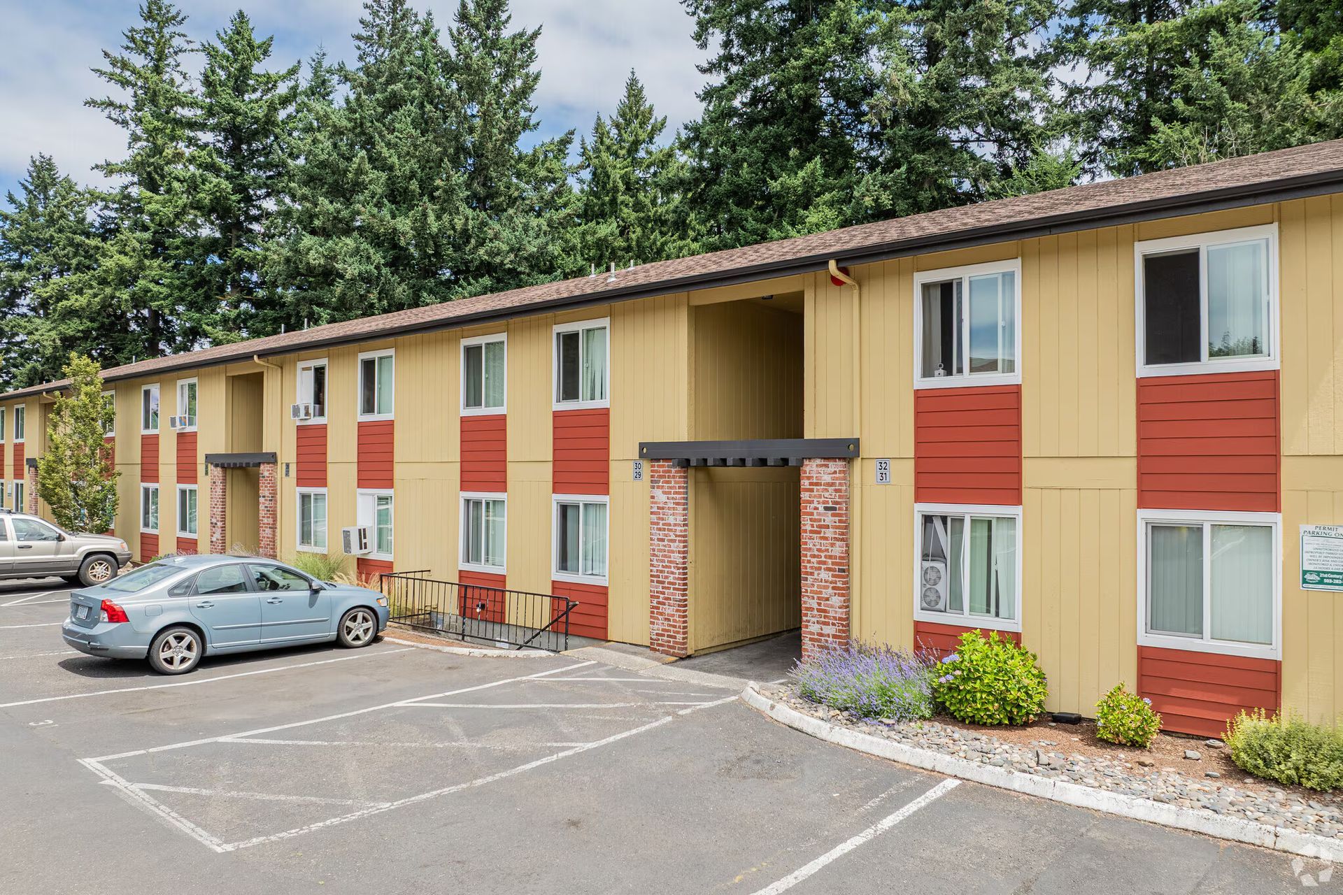Apartment building with beige and red exterior, parked car, and trees in front