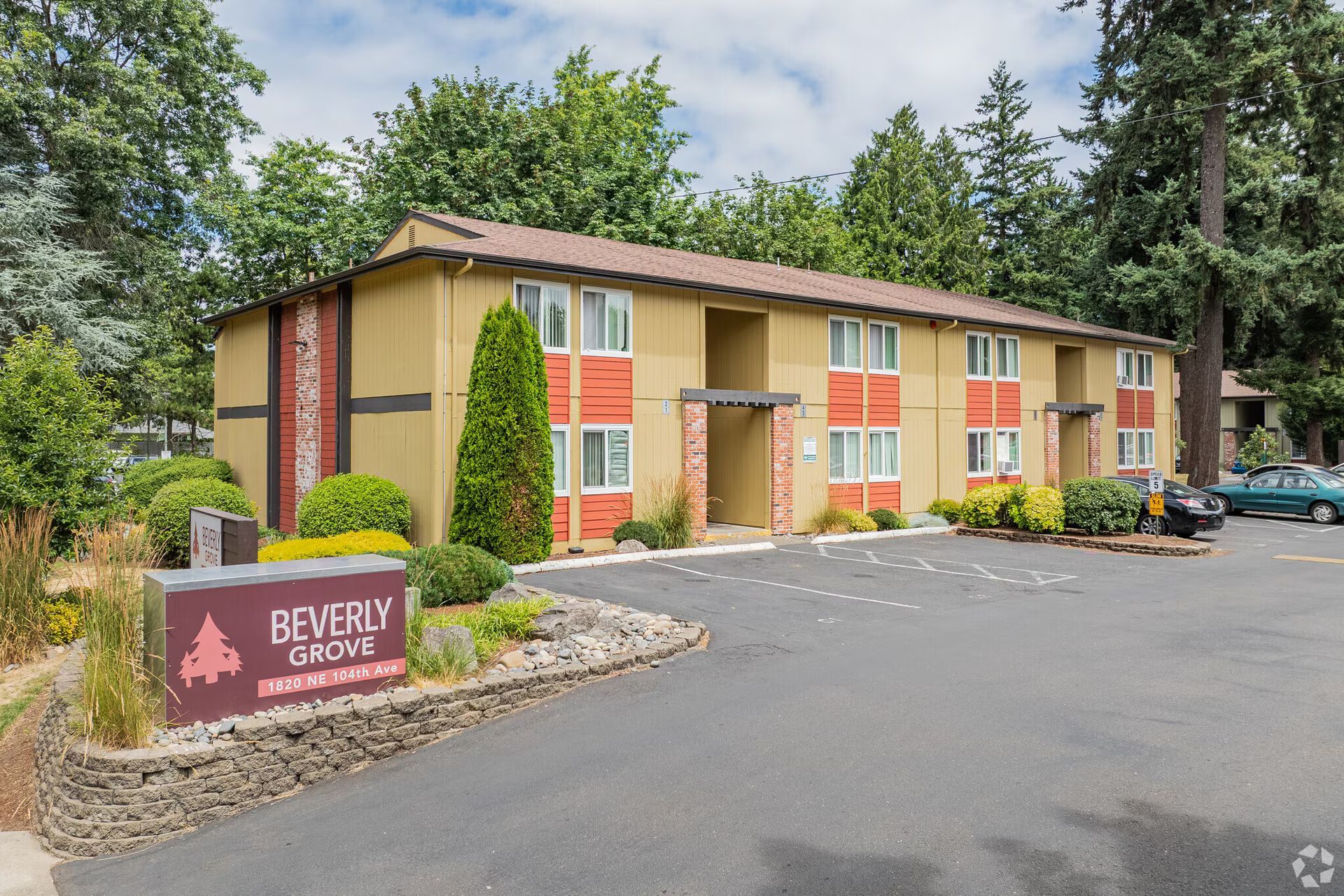 Apartment building exterior with a sign reading Beverly Gardens, surrounded by trees and parking lot.
