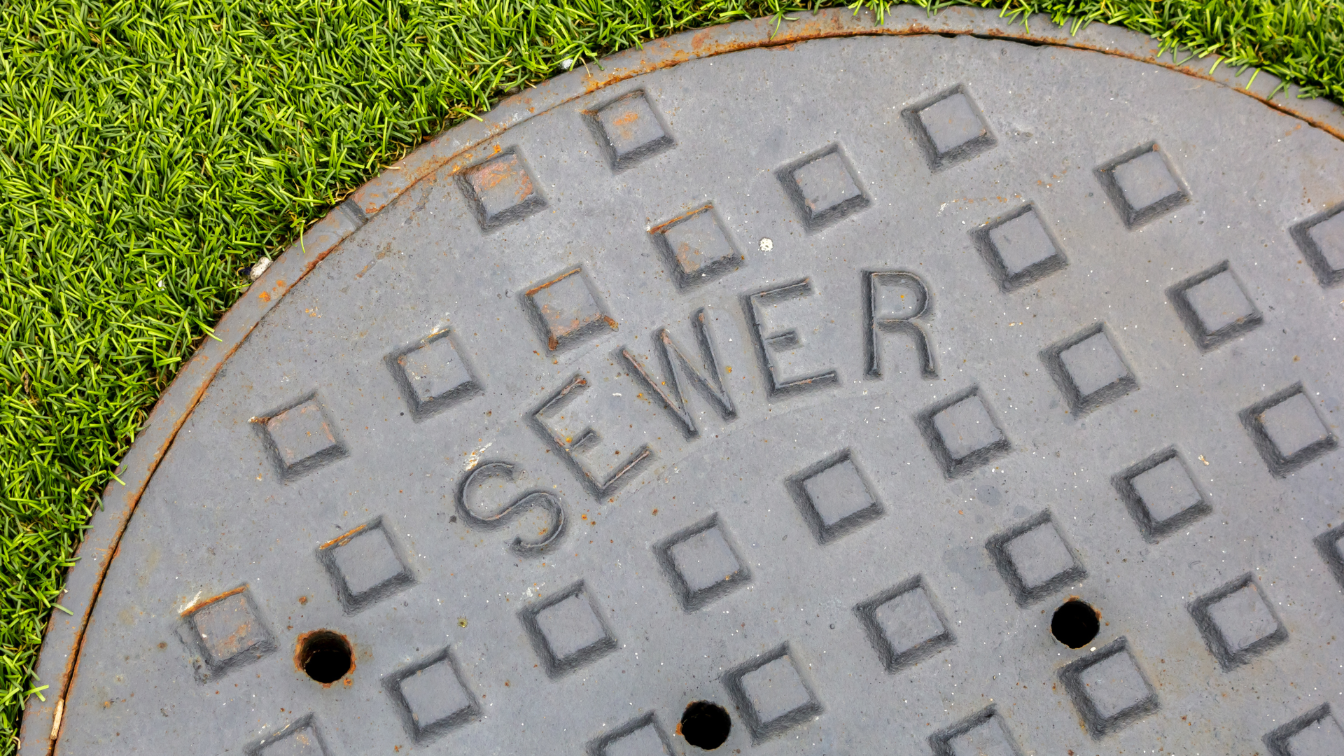 A round, gray metal sewer manhole cover with a grid pattern and the word