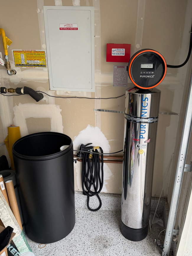 Utility closet with a black trash can, wall-mounted white panel, red alarm box, and shiny silver tank.