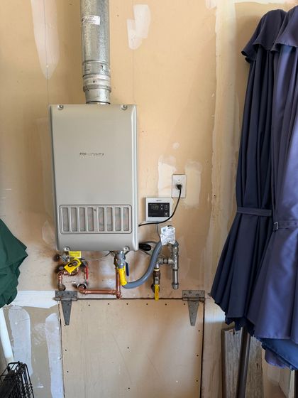 Water heater tank on a concrete platform in a garage, with laundry and cleaning items nearby.
