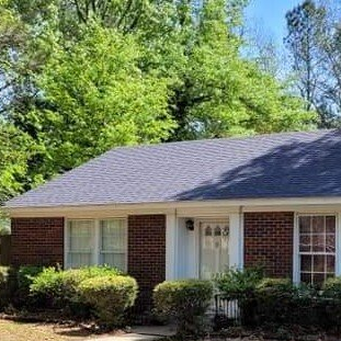 Single-story brick house with gray roof, white front door, and green trees in the yard