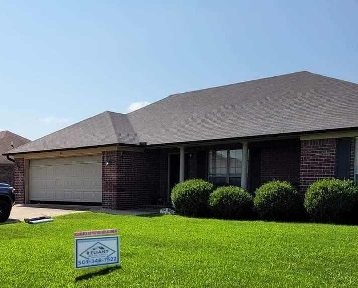 Single-story brick house with a gray roof, garage, and a for-sale sign on a green lawn.