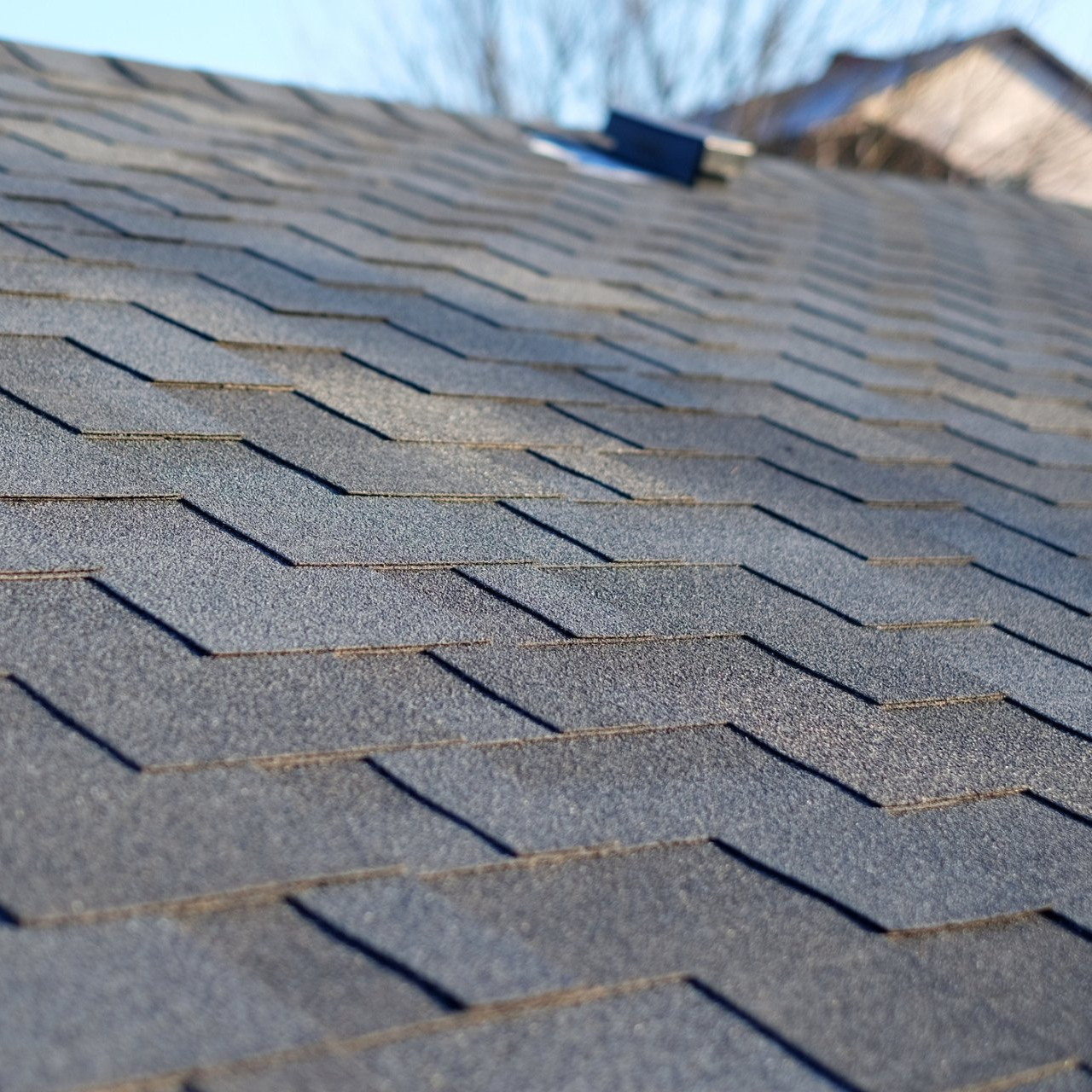 Close-up of gray asphalt shingles on a sloped roof with a blurred rooftop vent in the background